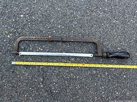 Hacksaw measuring about 18 inches in length on asphalt surface with visible wooden handle and metal frame.