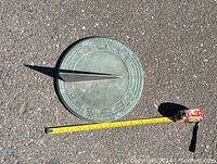 Antique metal sundial laying flat on asphalt next to a measuring tape, showing its dimensions.