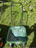 Front view of Scotts Turf Builder Mini fertilizer spreader on grass with plant backdrop, showing green hopper, black handle, and wheels.