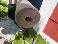 End view of a partially used roll of gray woven weed preventer fabric placed outdoors on a patio near plants.