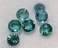 Seven round-cut genuine emerald gemstones laid out on a white surface, showing varying shades of green and natural inclusions.
