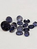 Photo showing a collection of 11 genuine iolite gemstones of various shapes and sizes, mostly oval and round cuts, with violet-blue coloring and faceted surfaces.