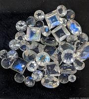 View of approx 32 loose genuine moonstone gemstones on a black background, showing various cuts including round, oval, and square with visible blue adularescence.