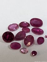 Collection of twelve faceted genuine rubies in various shapes and shades of red and pink against a white background.