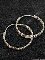 Pair of sterling silver twisted hoop earrings shown on black felt background, one earring overlapping the other, displaying twisted rope design and latch closure.