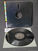 Black vinyl record with black label and white text, laid flat with original sleeve beneath it showing a black front cover with colorful stripes on left edge.