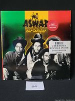 Front cover of Aswad vinyl record "Not Satisfied" showing band photo and colorful background with a sticker noting the free 10" single included.