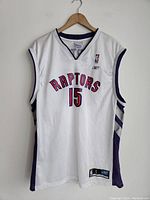 Front of white sleeveless Toronto Raptors basketball jersey with purple trim, number 15 on front, hanging on wooden hanger.
