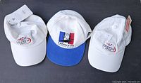 Three caps laid flat on a dark surface: two white US Open hats with tags, one showing staining, and a France 98 Soccer hat with a blue brim and multi-colored logo.