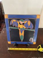 Front view of a square ceramic tile featuring a colorful parrot with wings spread, measuring about 7 inches with a tape measure in front for scale.