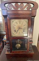 Front view of the antique wooden mantel clock showing carved top, glass front with pendulum, and aged clock face with Roman numerals.