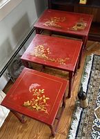 Three red Chinese nesting tables stacked with visible hand-painted gold designs on each top showing flowers and a pagoda.