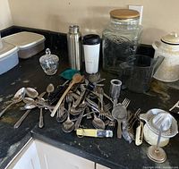 View of an assortment of kitchen utensils including spoons, forks, wooden spoons, a nutmeg grater, a yellow plastic pineapple corer, two travel mugs, plastic food containers, and glass jars.
