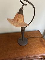 Full view of single table lamp showing curved metal arm and wide flared mottled glass shade in warm orange and amber. Base is round and metal with a dark patina finish.