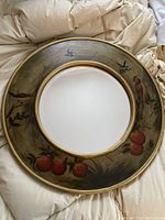 Round decorative mirror with painted design of apples and birds on the frame laid on a cloth surface.