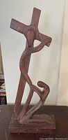 Front left angled view of the tall abstract wooden statue showing the figure carrying the cross and the rectangular base.