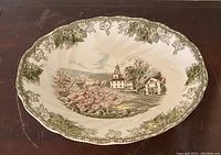 Front view of oval ceramic serving plate with detailed village scene and grapevine border design.