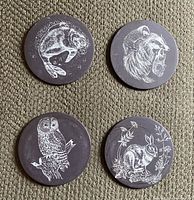 Four round slate coasters arranged on carpet, each engraved with different animal images - platypus, roaring bear, owl, and rabbit.