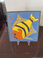 Front view of ceramic tile with colorful fish design displayed on a clear stand.