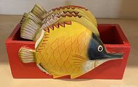 Front view showing five wooden fish-shaped coasters in yellow with orange scales, painted with black face detail, placed in a solid red rectangular wooden holder.