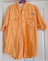 Front view of light orange Columbia button-up shirt hung on hanger, showing button details, two chest pockets, and collar.