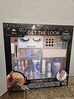 Front view of Disney Princess Cosmetic Kit in original packaging showing the items inside including eyelashes, eyeshadow, cream makeup tube, press-on nails, rhinestones, and cosmetic bag.