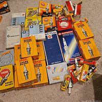 Overview photo showing multiple boxes of new vintage spark plugs mostly NGK brand with some AC Delco, Honda and Champion spark plugs visible