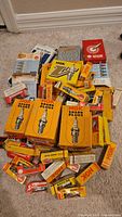 Photo showing a variety of spark plug boxes from NGK, Champion, and other brands spread on a carpeted floor.