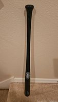 Full length photo of black Louisville Slugger baseball bat standing vertically against beige wall and carpet floor.