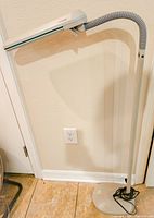 Full view of the white Ott-Lite floor lamp standing on tiled floor next to a wall outlet, showing adjustable neck and round metal base with cord wrapped.