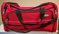 Front view of red Samsonite carry on bag with black handles and shoulder strap, showing front zippered pocket and oval metal Samsonite logo.