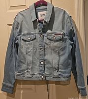 Front view of light blue Tommy Jeans denim jacket showing button front closure, collar, and chest pockets with button flaps.