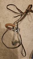 Full view of vintage western leather horse headstall with attached metal bit wrapped in a braided leather cover, laid flat on a carpeted surface.