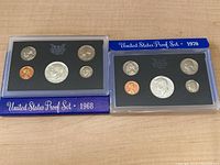 Two United States proof coin sets from 1968 and 1970 displayed side by side on a wood surface, each in plexiglass cases with original blue outer sleeves.