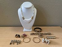 Full view of jewelry lot including white beaded necklace with pendant on display bust, three bracelets, several pairs of earrings, and brooch placed on table.