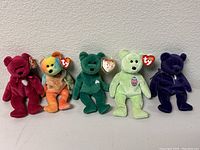 Five Beanie Babies standing in a row showing front view; includes Princess Diana bear, tie-dye bear, dark green shamrock bear, light green Erin bear, and dark purple velvet bear