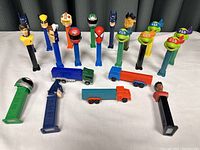 Group of 15 Pez dispensers arranged standing upright, featuring superhero and action figure characters, plus three small toy trucks.