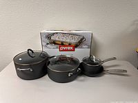 Group photo showing four Calphalon pots and saucepans with glass lids and stainless steel handles, along with boxed rectangular Pyrex baking dish.