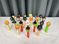 Group of 16 Halloween PEZ dispensers showing a variety of characters on colorful stems arranged on a white surface.