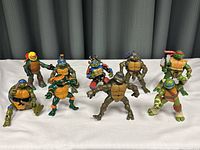 Group of nine Teenage Mutant Ninja Turtle action figures displayed in various fighting poses on a white cloth background against a dark curtain.