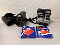 Yashica X30 camera with black case, Polaroid Sun 600 instant camera, and two boxes of Polaroid 600 film displayed on a white surface against a white wall.