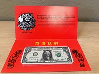 Front view of the uncirculated one dollar bill mounted on a red decorative card with Chinese gold characters and black illustrations, inside a protective sleeve.