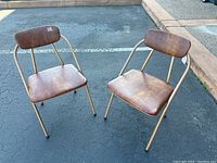 Pair of tan metal framed mid-century style folding chairs with brown padded seat and backrest, photographed from front angle on asphalt surface.