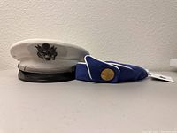 Photo of both hats side by side; left is the white captain's hat with silver eagle emblem and black visor; right is the blue Pan Am stewardess hat with white piping and gold Pan Am button.