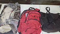 Three backpacks displayed on a white surface, from left to right: Terra 55 hiking backpack, red multi-compartment backpack, and black sling backpack.