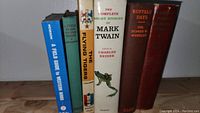 Seven hardcover books standing upright on a wooden surface, titles mostly relating to Mark Twain and western themes.