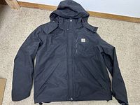 Black men's Carhartt Storm Defender jacket laid flat showing front with hood and chest logo patch.