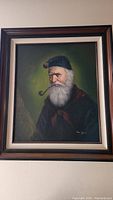 Framed oil painting of an old man with white beard smoking a pipe, shown hanging on a wall.