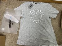 Top-down view of new gray Gymshark t-shirt with white fitness logo and shark graphic, laid flat on carpet with plastic Gymshark XL bag beside it.