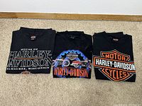 Front views of three black Harley Davidson short-sleeve t-shirts, showing various logo graphics including 'House of Harley-Davidson Milwaukee, Wisconsin', a motorcycle graphic, and the orange Harley-Davidson Cycle logo.